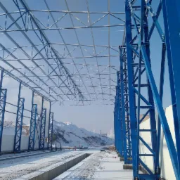 Konstrukcja stalowa hali w trakcie budowy, widoczne niebieskie słupy i kratownica dachu, na ziemi śnieg, w tle ośnieżone wzgórza.