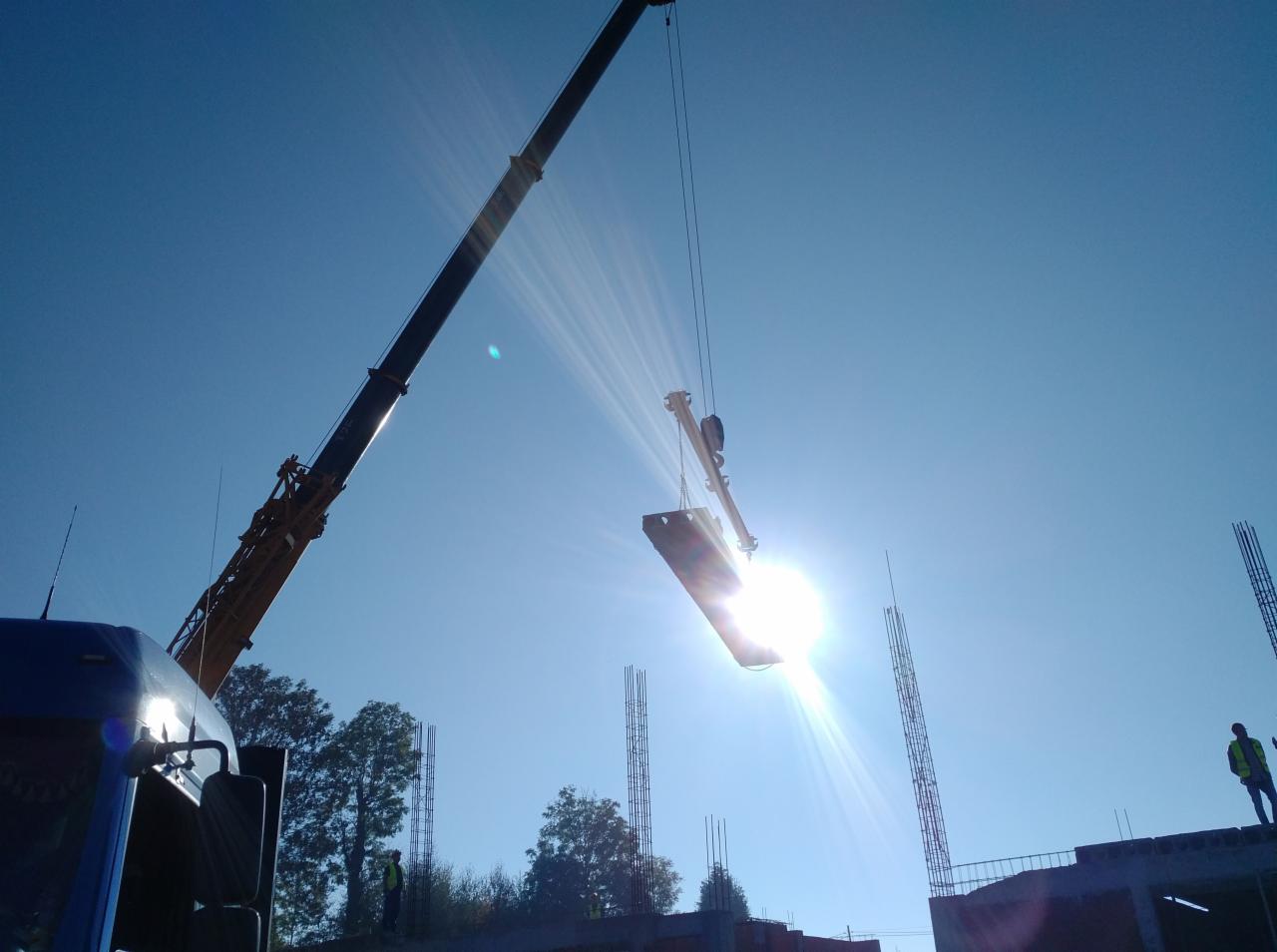 Żuraw przenosi prefabrykowany element betonowy na placu budowy, widoczne zbrojenia i pracownik w kamizelce odblaskowej na tle słonecznego nieba.