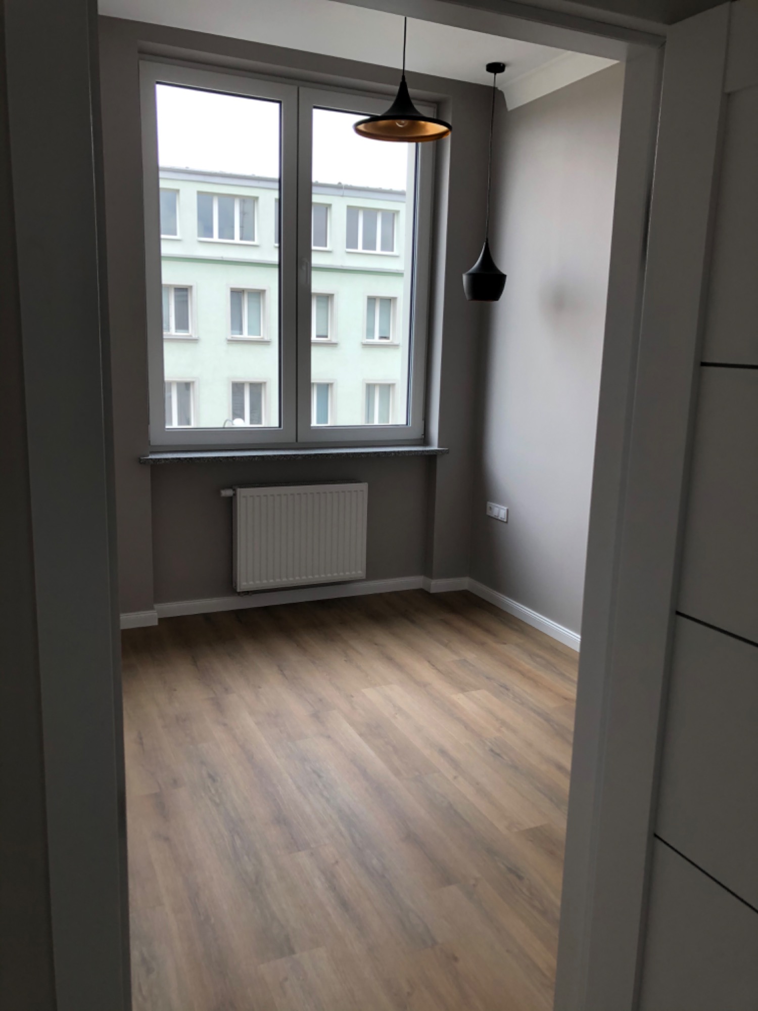 Pusty pokój po remoncie z panelami podłogowymi w odcieniach brązu, szarą ścianą z grzejnikiem pod oknem i dwiema wiszącymi lampami w stylu retro.