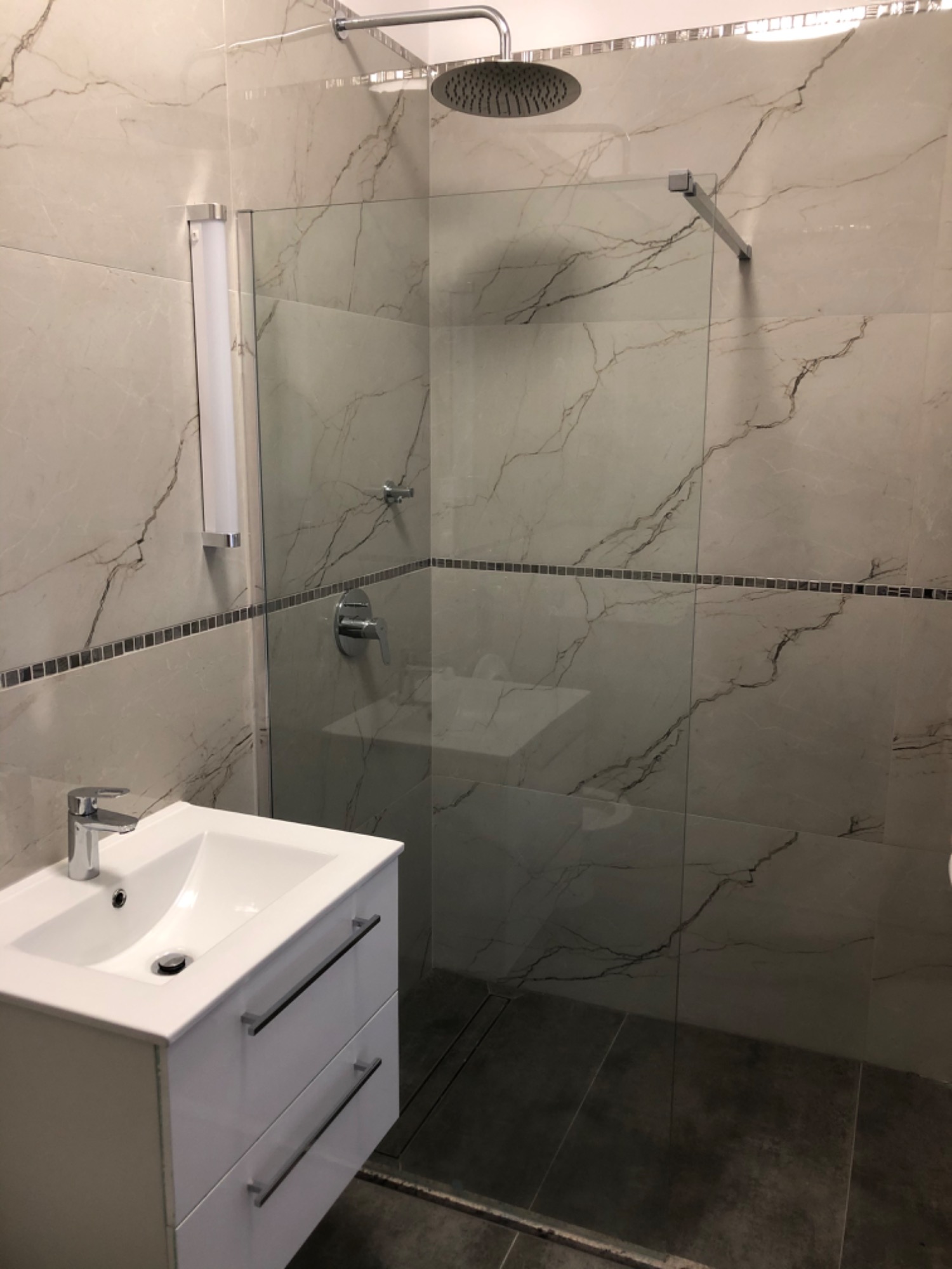 Nowoczesna łazienka z marmurowymi płytkami, szklanym panelem prysznicowym typu walk-in, białą szafką pod umywalką z chromowanymi uchwytami i deszczownicą.