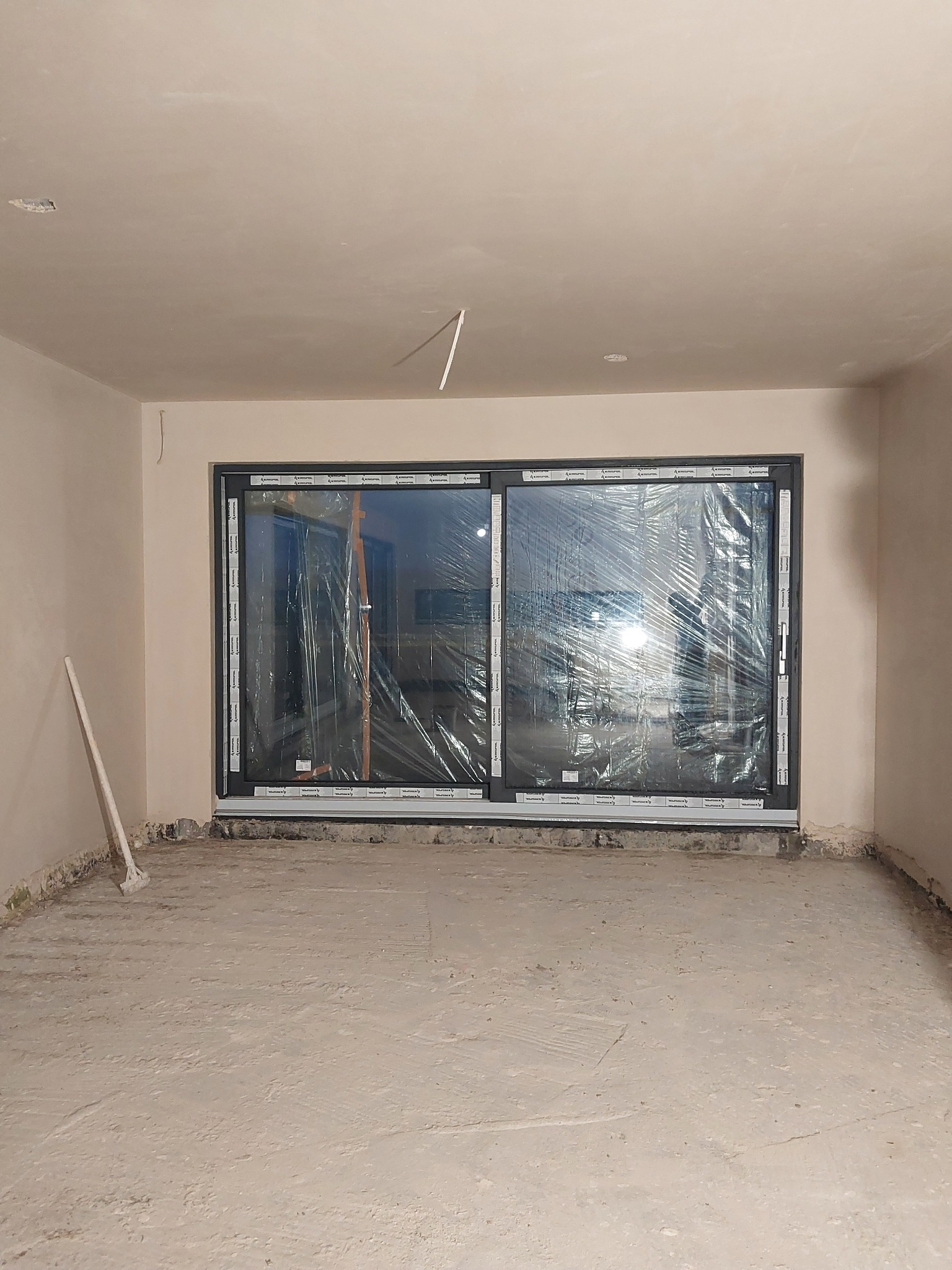 Pokój w trakcie prac wykończeniowych: ściany po nałożeniu gładzi, okno przesuwne zabezpieczone folią, surowa wylewka na podłodze, widoczna szpachelka oparta o ścianę.