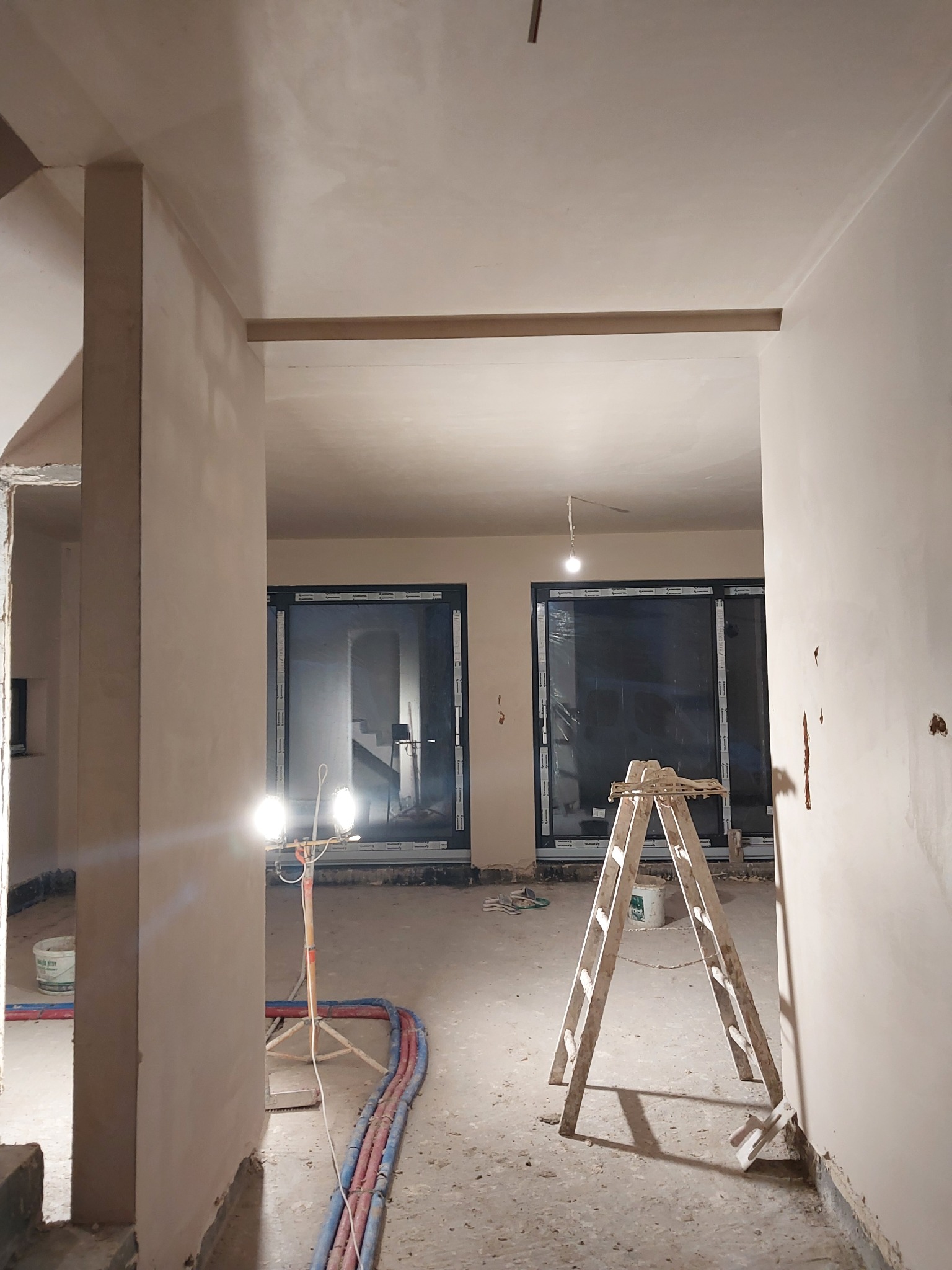 Wnętrze w trakcie prac wykończeniowych: świeżo otynkowane ściany, drabina oparta o ścianę, lampa budowlana oświetlająca pomieszczenie, widoczne przewody instalacyjne na podłodze.