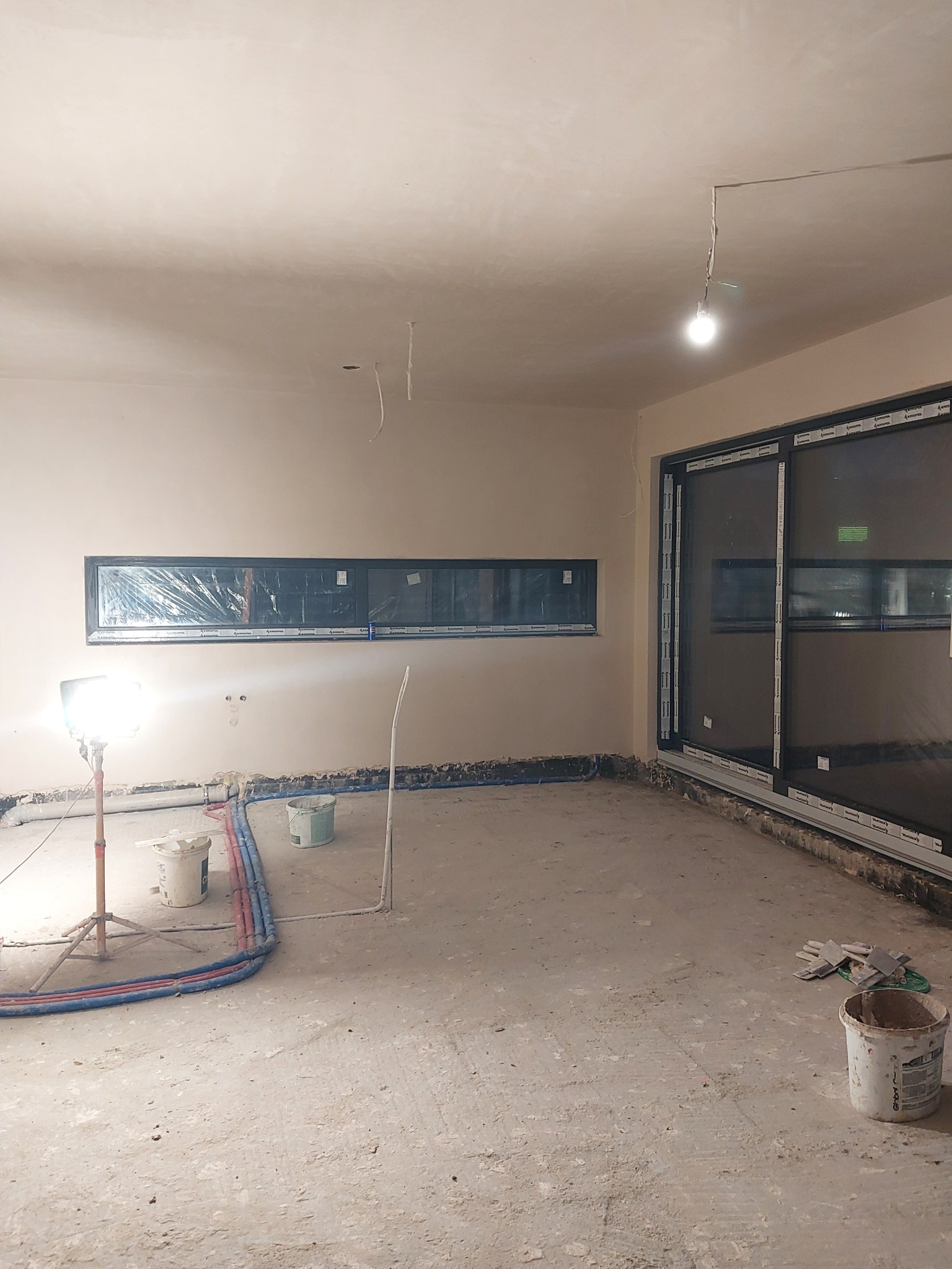 Świeżo otynkowane ściany w pomieszczeniu, z widocznymi przewodami instalacji grzewczej na podłodze, okna zabezpieczone folią, lampa budowlana oświetla wnętrze, widoczne narzędzia i wiaderka po farbie.