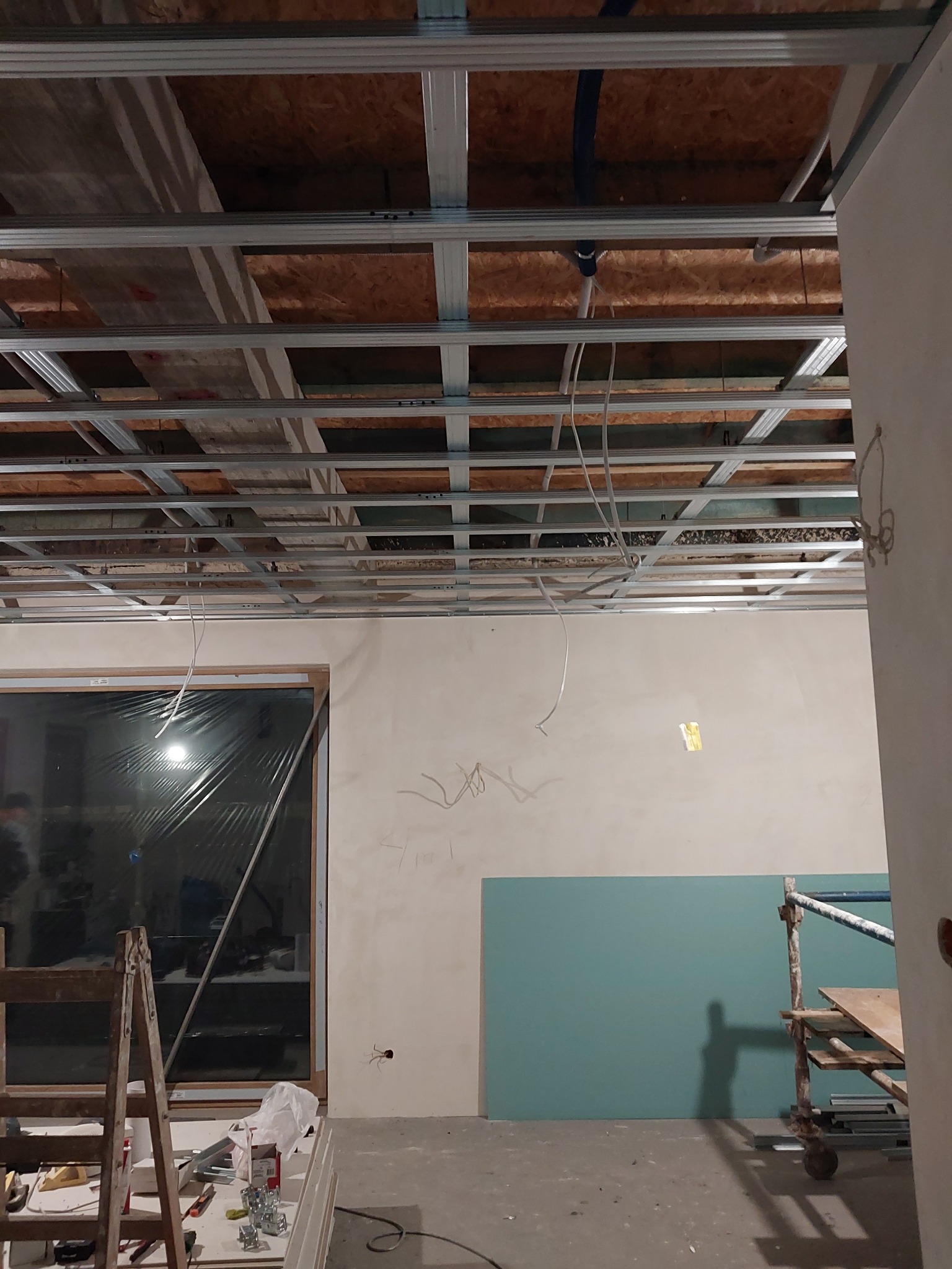 Montaż sufitu podwieszanego z płyt gipsowo-kartonowych, widoczne profile metalowe, instalacje oraz częściowo otynkowana ściana z oknem i drabina.
