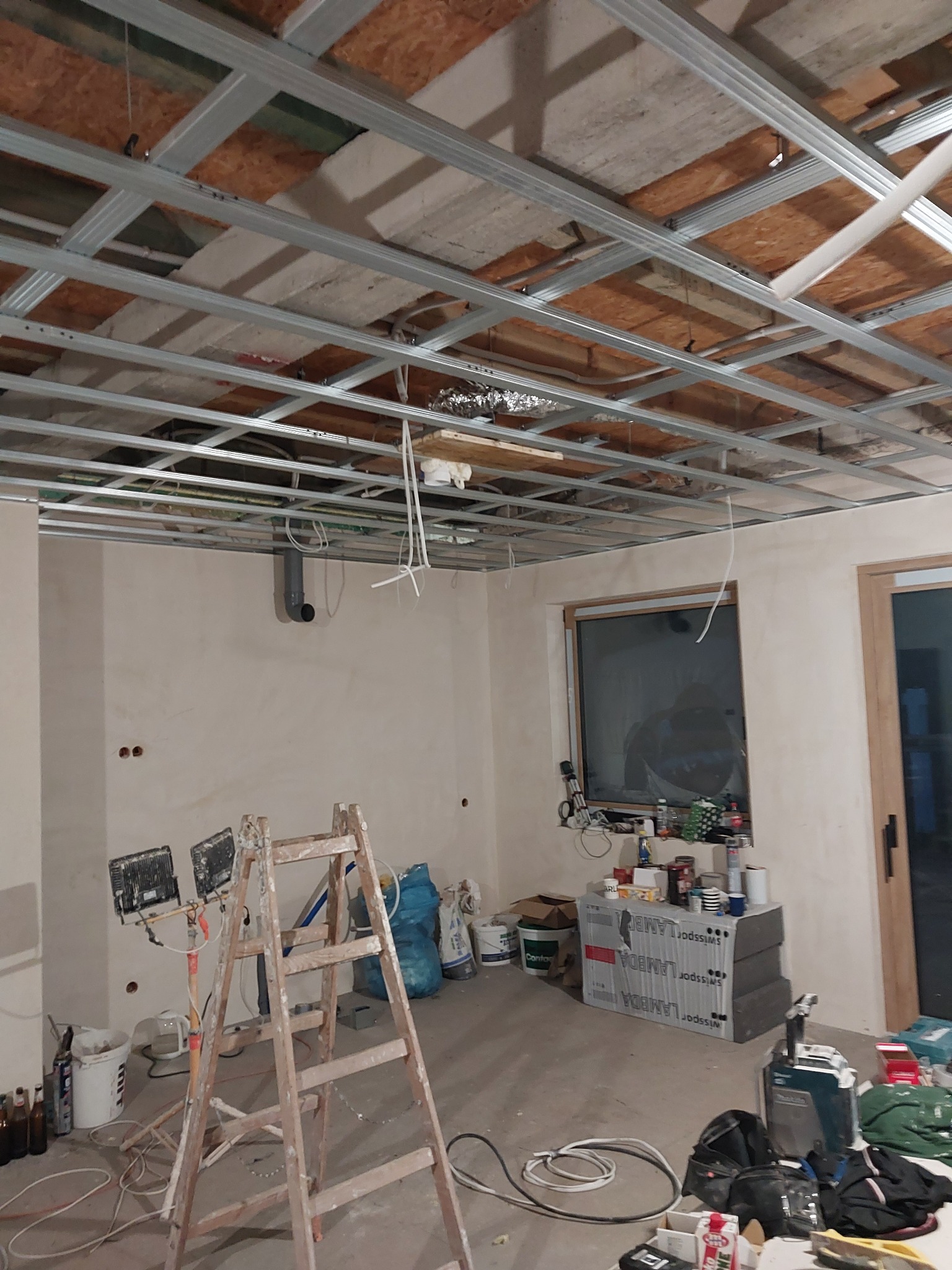 Wnętrze w trakcie montażu sufitu podwieszanego z metalowej konstrukcji, widoczne narzędzia, drabina i materiały budowlane.