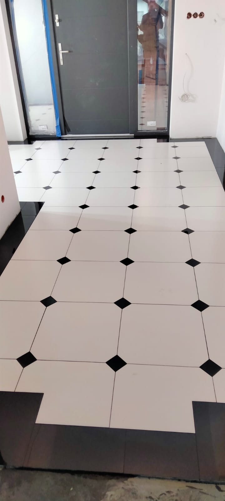 Ułożona podłoga z białych płytek z czarnymi rombami na skos, widoczny fragment czarnej listwy przypodłogowej, w tle szare drzwi z przeszkleniem i wystające przewody elektryczne.