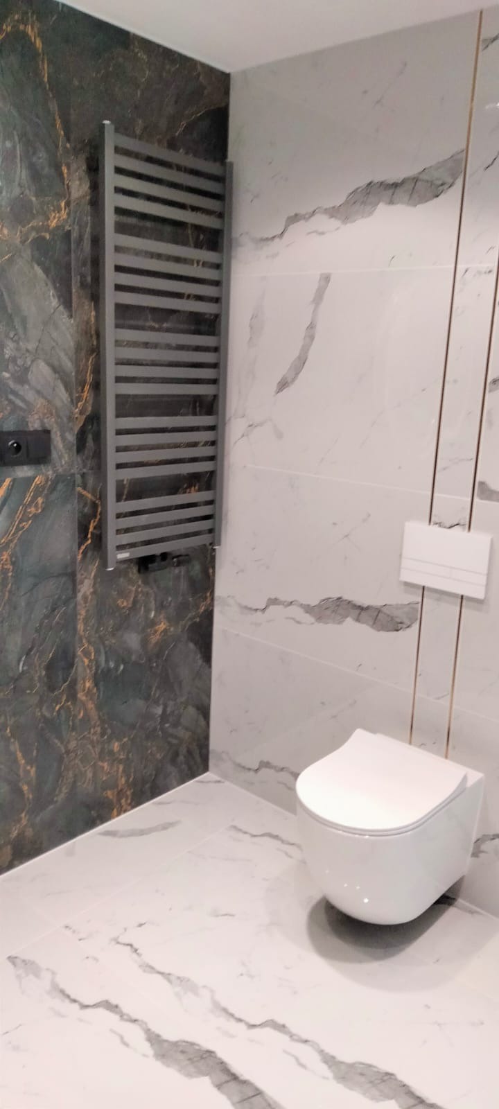 Nowoczesna łazienka z marmurowymi płytkami w odcieniach bieli i szarości, grzejnik drabinkowy w kolorze szarym na tle ciemnej ściany z żyłkowaniem w kolorze złota, wisząca toaleta.