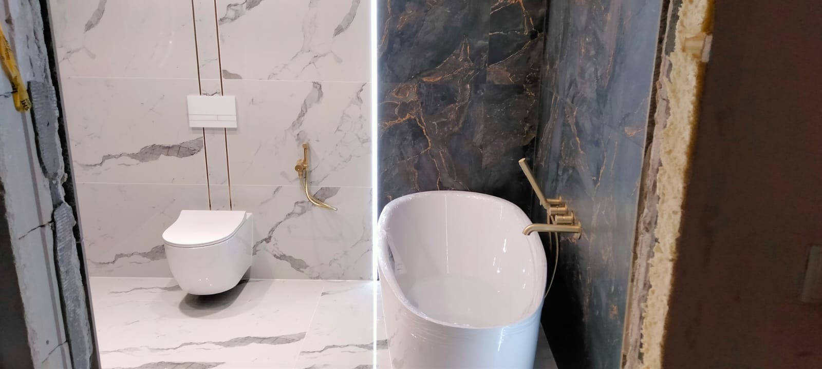 Nowoczesna łazienka w trakcie wykańczania: biała ceramika sanitarna (toaleta wisząca, wanna wolnostojąca w folii) na tle marmurowych płytek z złotymi akcentami, widoczne niedokończone obrzeża ścian.