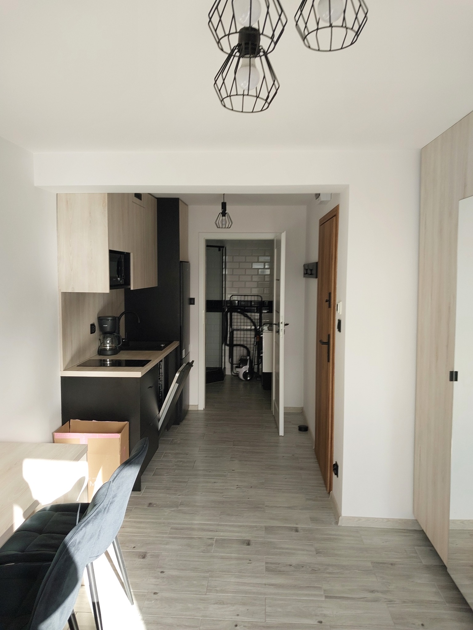 Nowoczesne wnętrze mieszkania z aneksem kuchennym w odcieniach bieli, szarości i czerni, z widocznymi lampami sufitowymi w stylu industrialnym i szarymi płytkami podłogowymi imitującymi drewno.