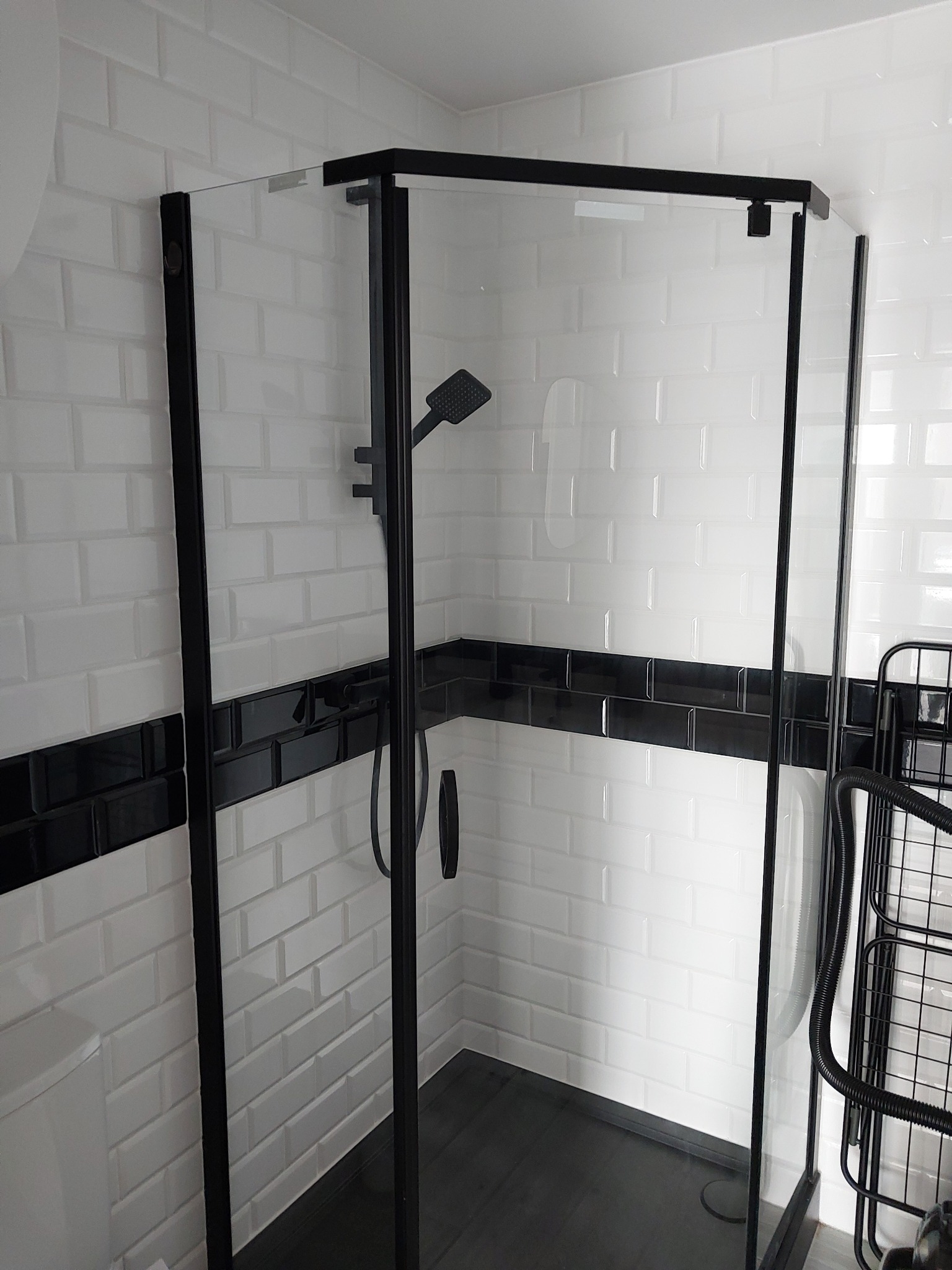 Nowoczesna kabina prysznicowa z czarną ramą i armaturą na tle ściany z białych, prostokątnych płytek z czarnym pasem dekoracyjnym.