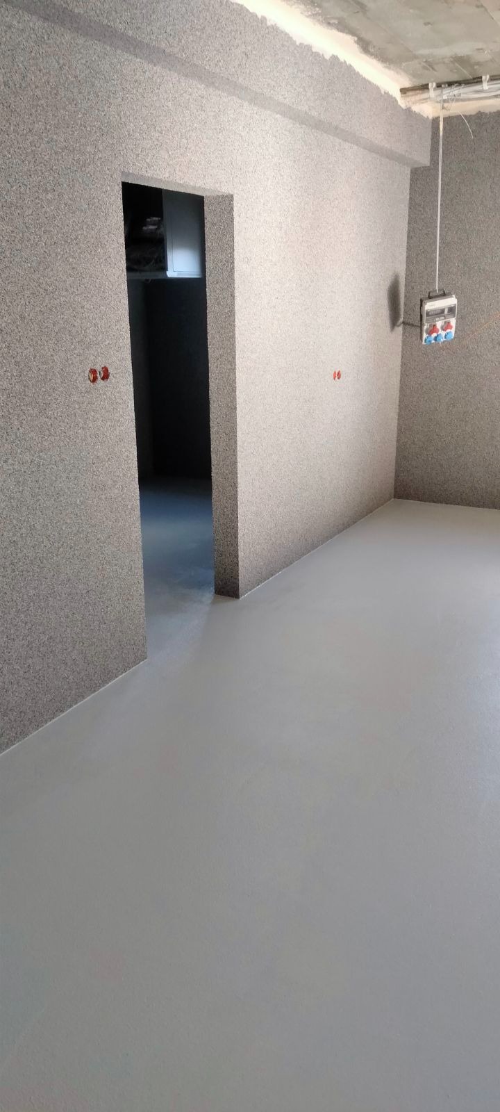 Pokój po nałożeniu tynku strukturalnego na ściany i wylaniu szarej posadzki samopoziomującej; widoczna instalacja elektryczna.