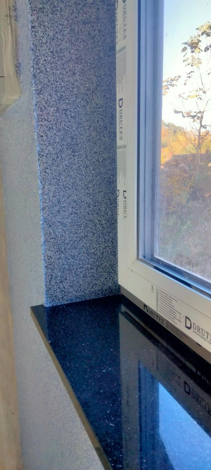 Szorstka, niebiesko-szara ściana z tynkiem strukturalnym obok okna z czarnym, błyszczącym parapetem z widokiem na drzewa w oddali.