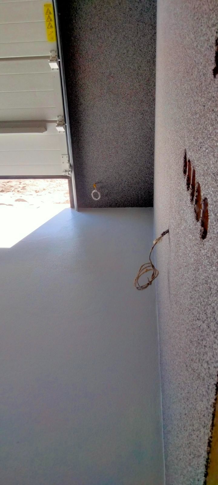 Ściana z nałożonym tynkiem strukturalnym, widoczne otwory pod gniazdka elektryczne i wystające przewody, brama garażowa w tle.