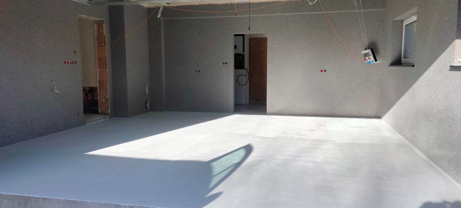 Wnętrze w stanie deweloperskim z nałożonym tynkiem maszynowym o fakturze baranka, jasna wylewka podłogowa, widoczny rozdzielacz elektryczny na ścianie i fragment okna.