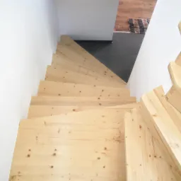 Widok z góry na jasne, drewniane schody spiralne, przy białej ścianie, z fragmentem szarej posadzki i dywanika widocznym na górnym poziomie.