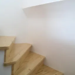 Drewniane schody z jasnego drewna, widoczne od dołu, z charakterystycznym usłojeniem, przylegające do białej ściany.