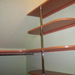 Półki z płyty wiórowej w kolorze jasnego drewna, zamocowane na metalowych rurach, dopasowane do skośnej ściany na poddaszu, widok z dołu.