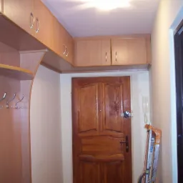 Przedpokój z drewnianą zabudową: szafki wiszące nad drzwiami, wieszak z półką i haczykami, widoczna drabina oparta o ścianę.