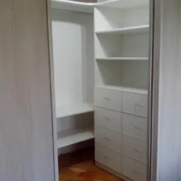 Wnętrze szafy wnękowej z białymi półkami i szufladami, widoczne z boku, z przesuwne drzwi częściowo otwarte, na podłodze parkiet w jodełkę.