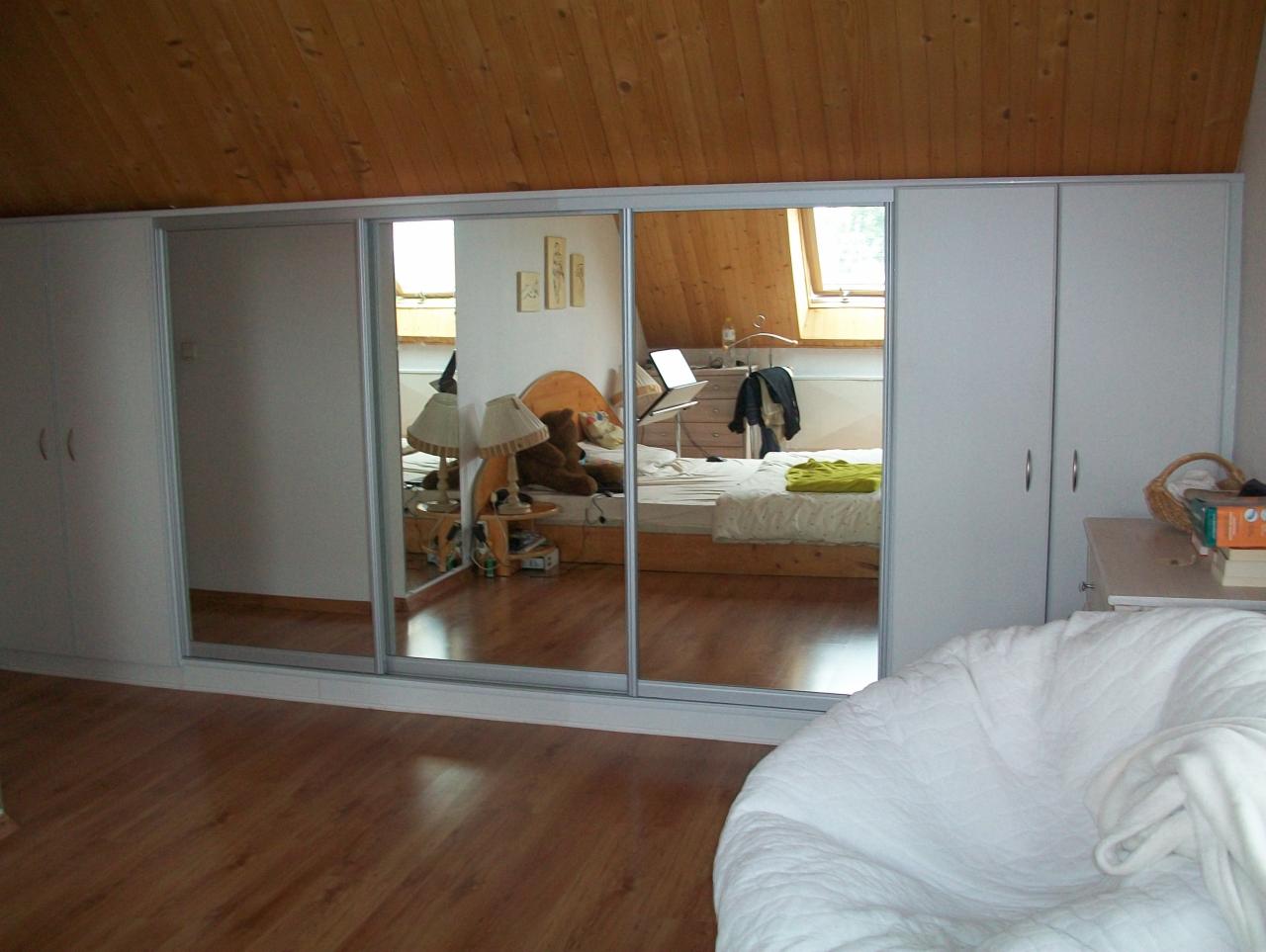 Biała szafa przesuwna z lustrami w sypialni na poddaszu, odbijająca widok łóżka, biurka i skośnego okna dachowego.