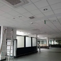 Sufit kasetonowy w jasnym pomieszczeniu biurowym z widoczną drabiną, kratkami wentylacyjnymi i oświetleniem punktowym.