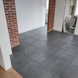 Wnętrze w trakcie prac wykończeniowych: szare płytki podłogowe ułożone w kwadratową siatkę, białe ściany z fragmentami surowej cegły, widoczne ślady butów na podłodze oraz wiadro z klejem malarskim...