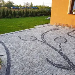 Podjazd z szarej nawierzchni ułożonej w motyw roślinny z ciemniejszych elementów, widok na dom z pomarańczową elewacją, trawnik i rząd iglaków.