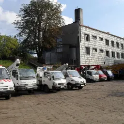 Rząd zaparkowanych samochodów dostawczych z podnośnikami koszowymi na placu, na tle szarego budynku z licznymi oknami i drzewami.