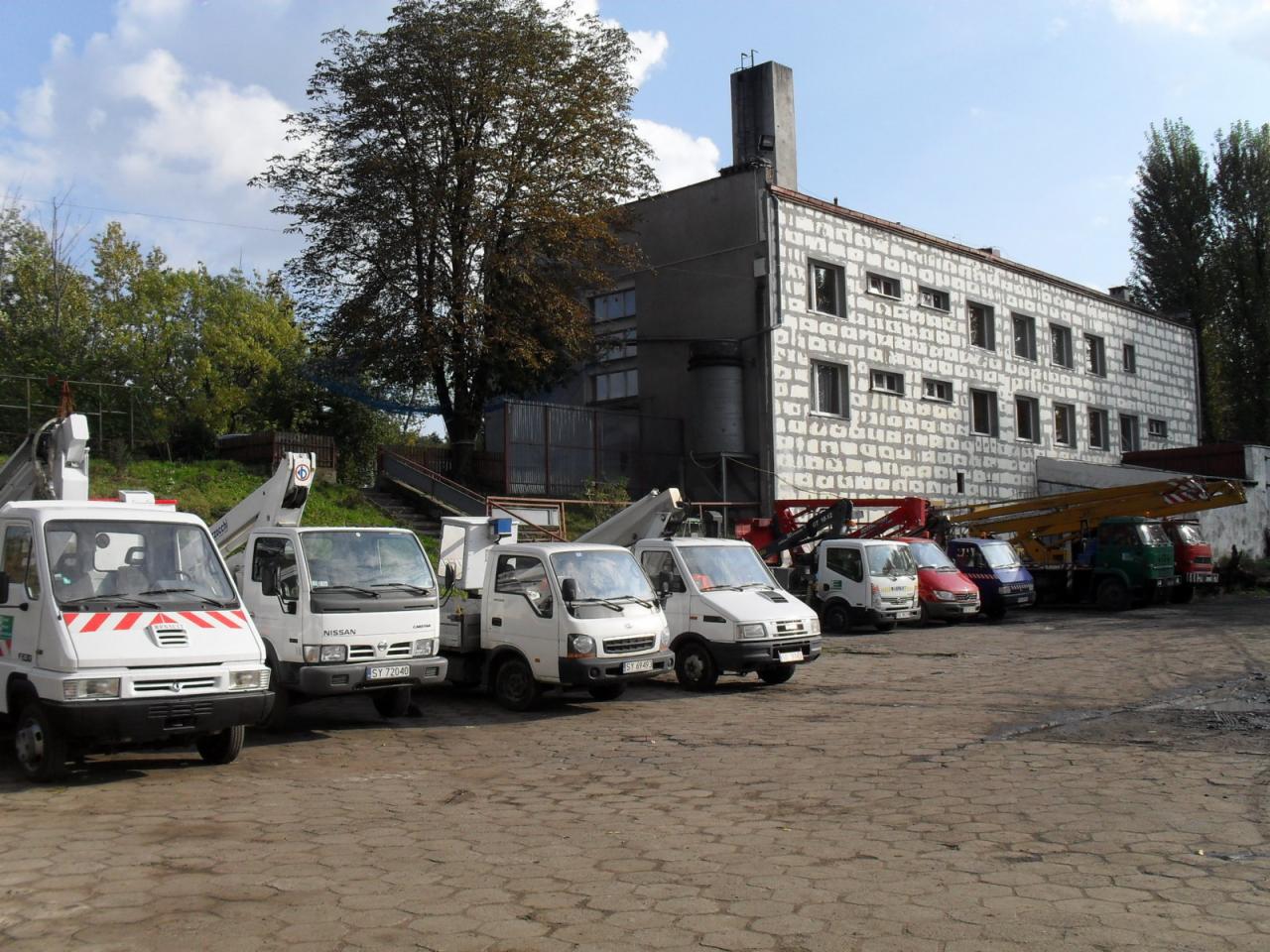 Rząd zaparkowanych samochodów dostawczych z podnośnikami koszowymi na placu, na tle szarego budynku z licznymi oknami i drzewami.