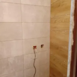 Ściana łazienki wyłożona jasnoszarymi, poziomymi płytkami z fugami oraz fragment ściany z płytkami imitującymi drewno; widoczne puszki elektryczne z wystającymi przewodami i plastikowe krzyżyki...