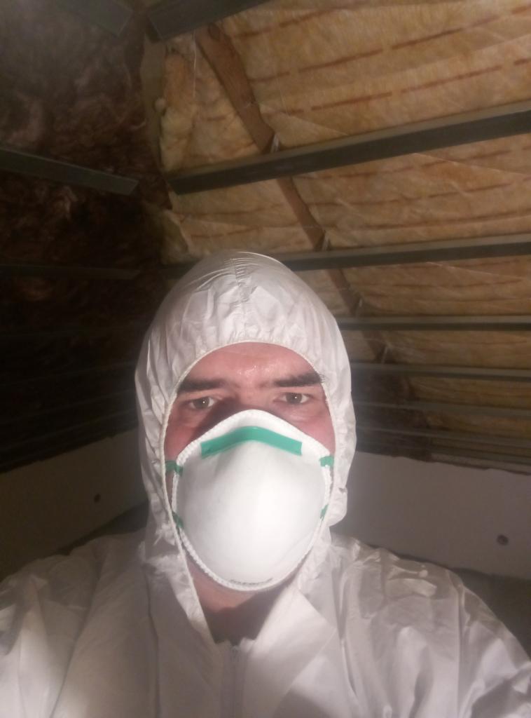 Pracownik w kombinezonie ochronnym i masce, instalujący izolację w poddaszu z drewnianą konstrukcją i metalowymi profilami.