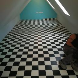 F.U.H. SAKS - Poddasze z ukończoną podłogą w czarno-białą szachownicę, widoczny fragment osoby pracującej przy krawędzi, niebieska ściana szczytowa z gniazdkami elektrycznymi.