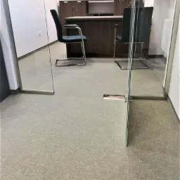 Biuro w Gdyni z szarą wykładziną dywanową o delikatnym wzorze, widok przez szklane drzwi, krzesła biurowe i zabudowa meblowa w tle.