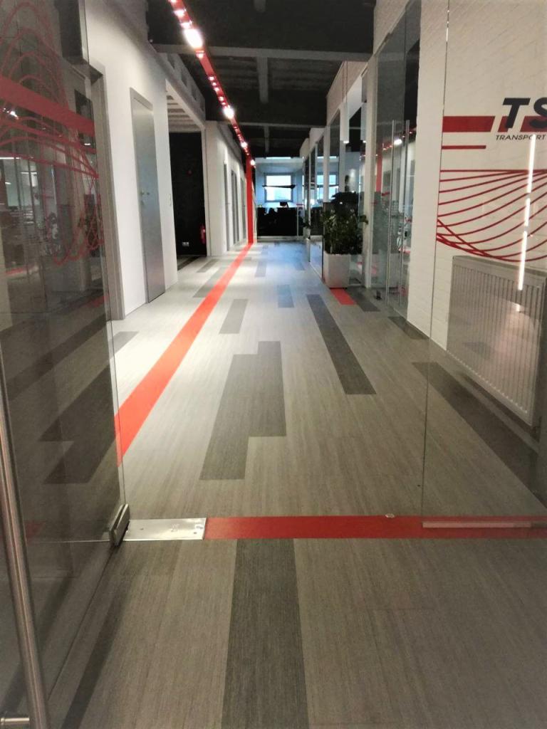 Długi korytarz biurowy z podłogą w odcieniach szarości z geometrycznym wzorem i czerwoną linią akcentującą, widok zza szklanych drzwi.