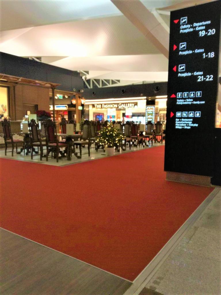 Czerwona podłoga z wykładziny w przestrzeni publicznej, prawdopodobnie lotnisko, z widocznymi stolikami i krzesłami w tle oraz tablicą informacyjną z oznaczeniami bramek.