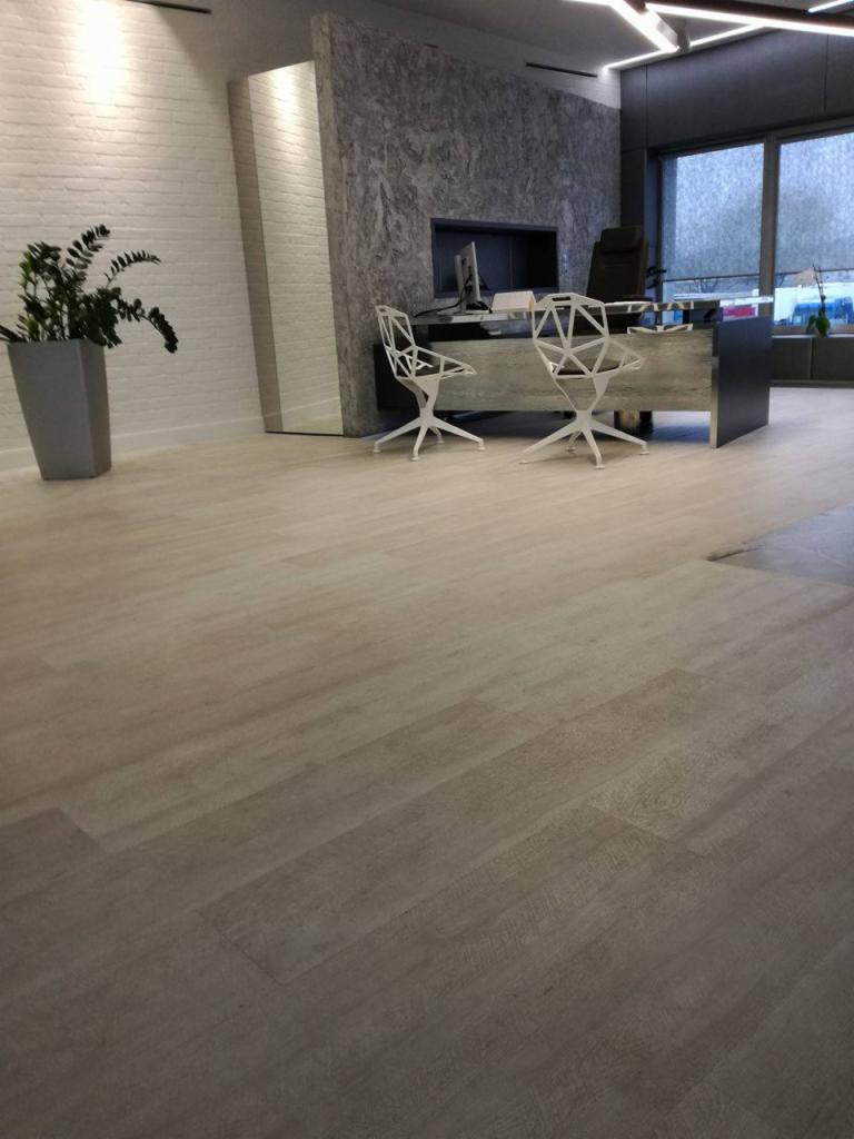 Jasna, drewnopodobna podłoga w nowoczesnym biurze z białej cegły i szarym akcentem, widoczne geometryczne krzesła i biurko.