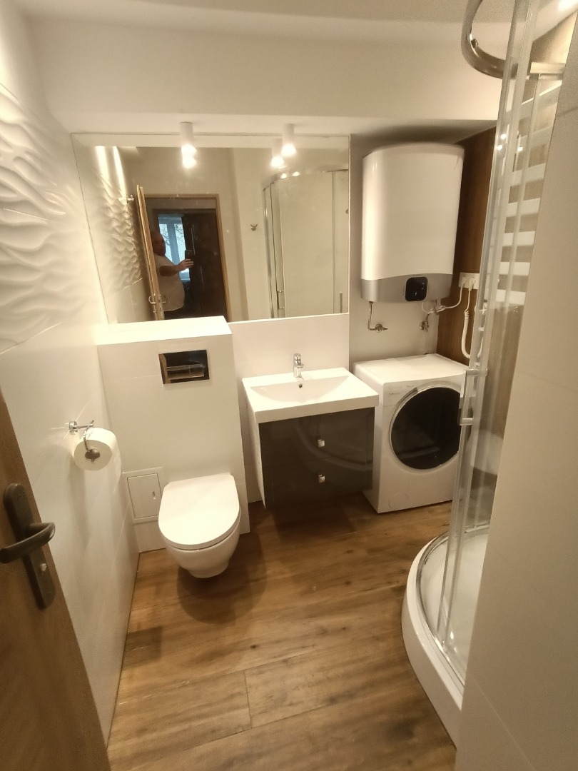 Nowoczesna łazienka z drewnopodobną podłogą, białą armaturą, kabiną prysznicową z przezroczystego szkła, umywalką z szafką w kolorze grafitowym, podwieszaną toaletą i pralką.