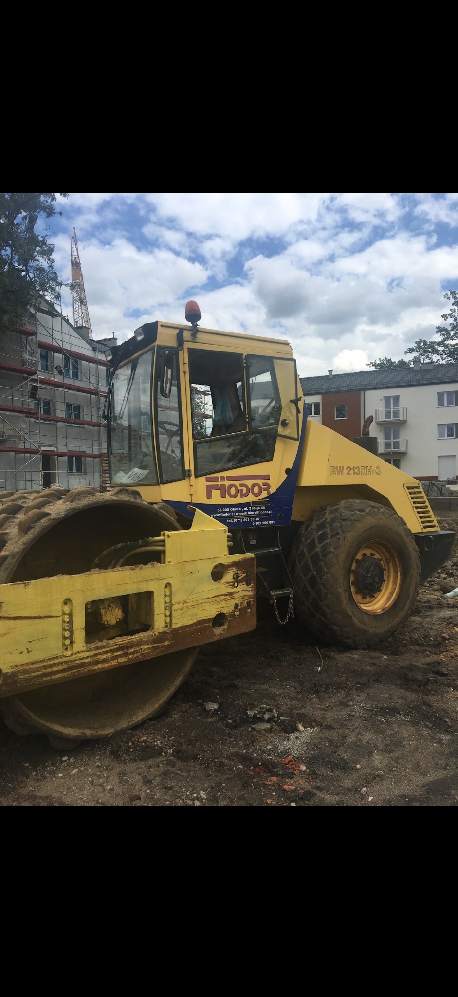 Żółty walec drogowy marki Fiodor BW 213DH-3 na placu budowy, w tle budynek w rusztowaniach i żuraw. Sprzęt budowlany do robót ziemnych.