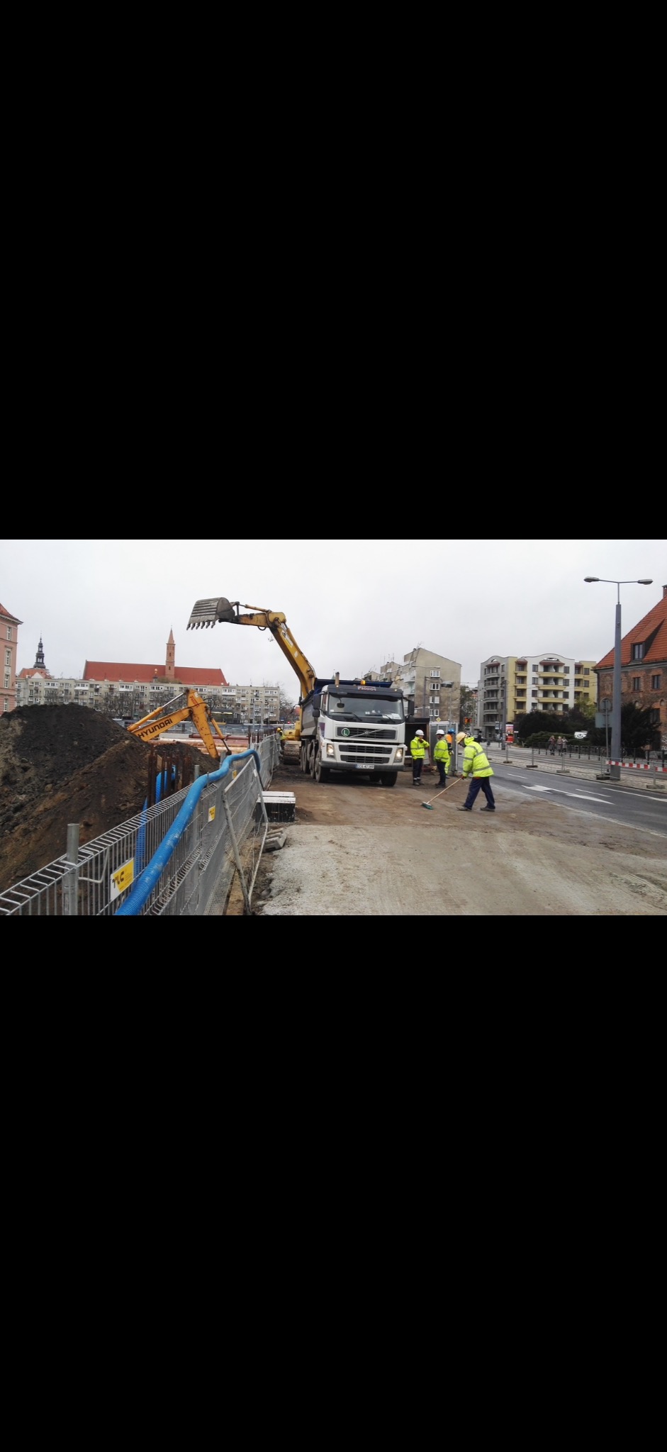 Żółty koparkoładowarka Hyundai ładuje ziemię na ciężarówkę. W tle budynki miejskie i pracownicy w odblaskowych kamizelkach. Prace ziemne w toku.