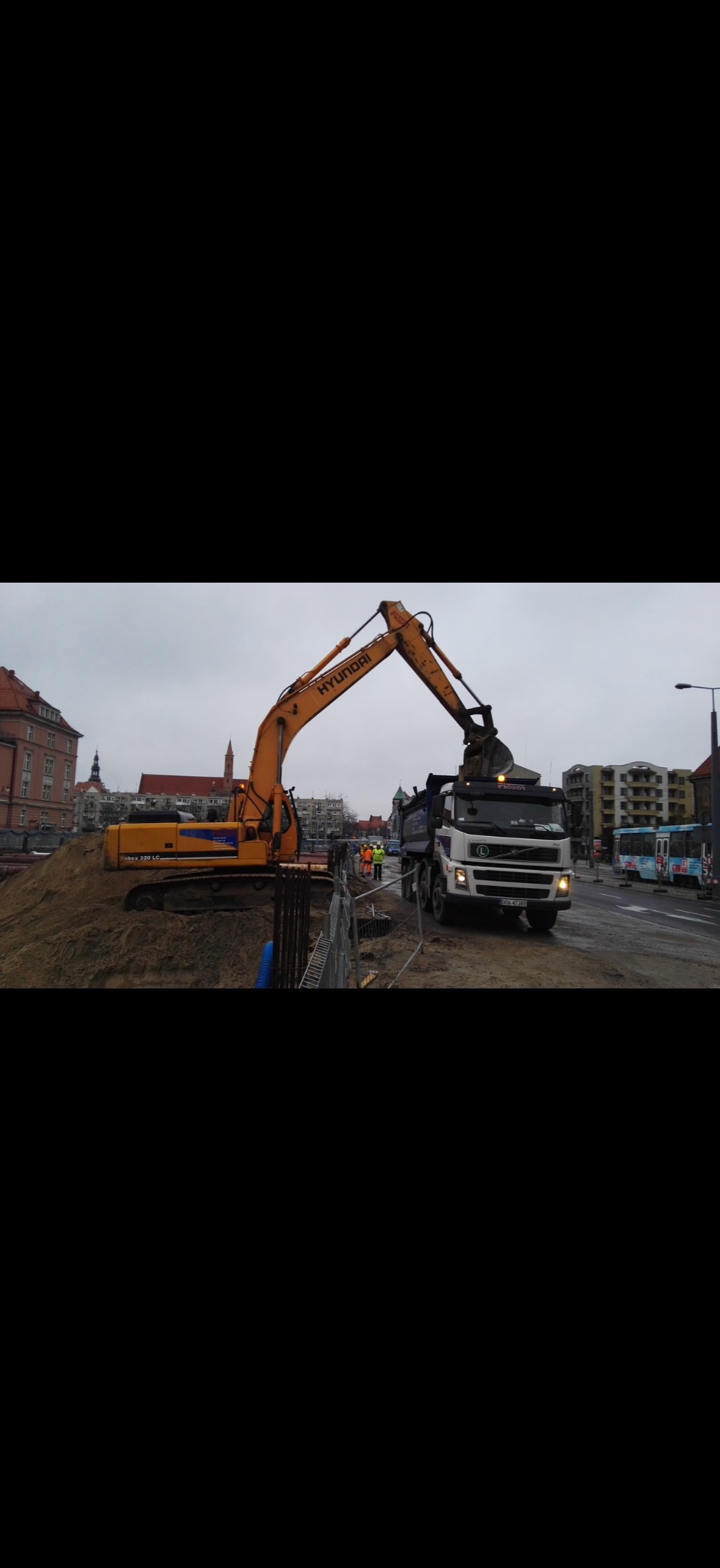 Żółta koparka Hyundai nabiera ziemię na ciężarówkę na tle miejskiego krajobrazu z budynkami i przejeżdżającym autobusem. Widoczni pracownicy w odblaskowych kamizelkach.