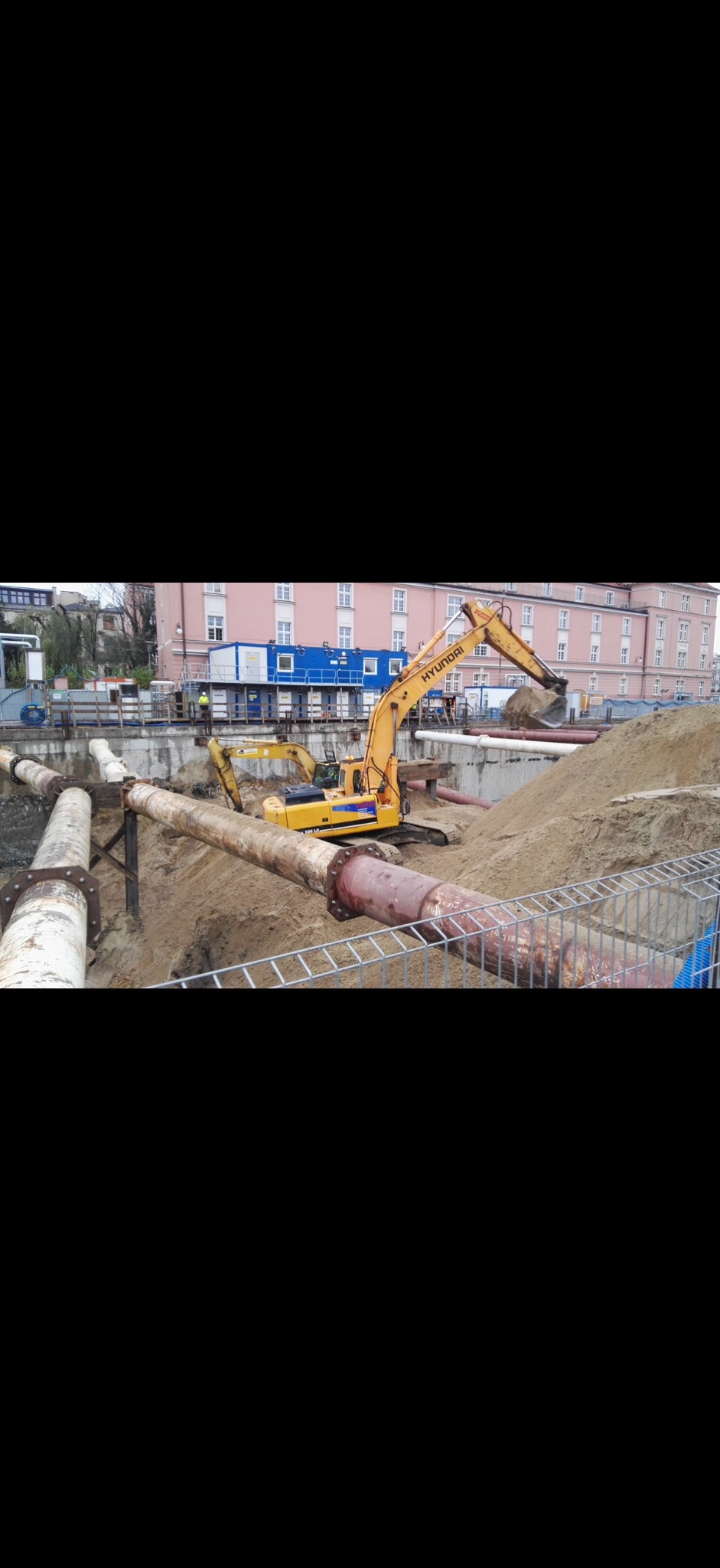 Żółta koparka Hyundai w wykopie, obok rury ciepłownicze. W tle budynek i kontener biurowy. Prace ziemne w trakcie realizacji.