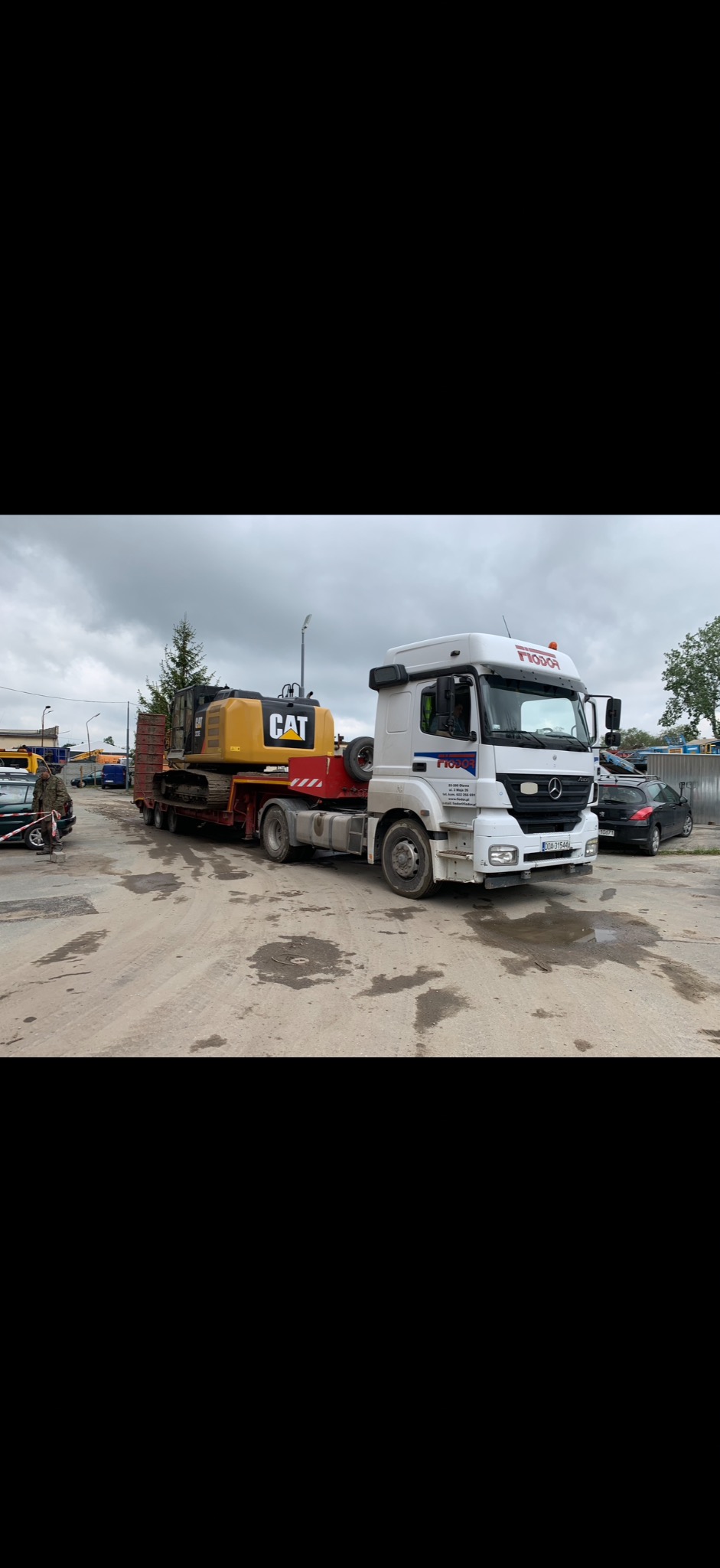 Transport niskopodwoziowy: biała ciężarówka Mercedes przewozi żółtą koparkę CAT na czerwonej platformie. Widoczne logo firmy transportowej na kabinie.