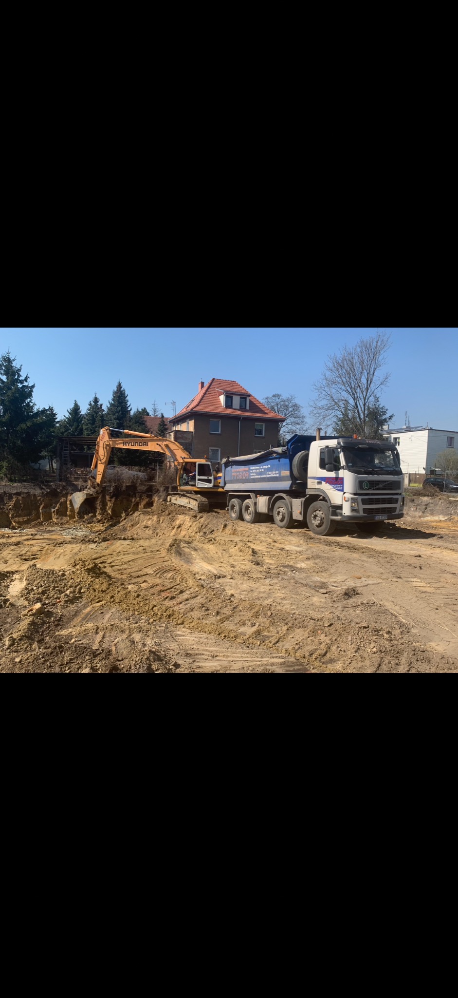 Żółta koparka Hyundai i ciężarówka Volvo na placu budowy, wykop pod fundamenty domu, w tle budynek z czerwoną dachówką i drzewa. Słoneczny dzień.