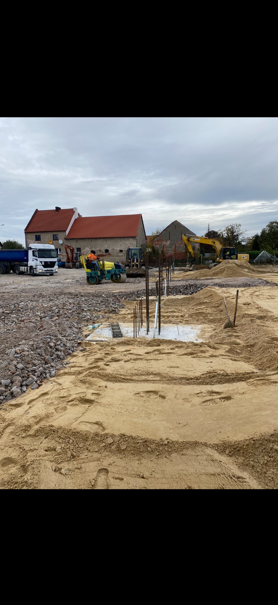 Przygotowanie terenu pod budowę: wyrównany piasek, wystające pręty zbrojeniowe, w tle maszyny budowlane i budynki. Widok na plac budowy w pochmurny dzień.