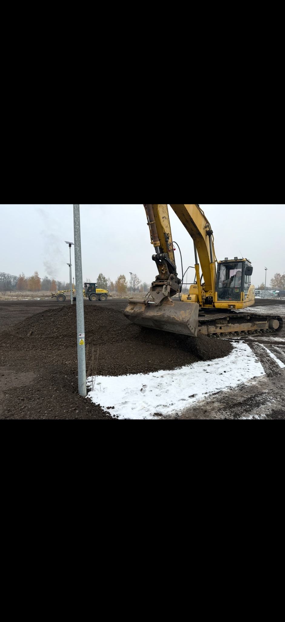 Żółta koparka nabiera ziemię na pryzmę. W tle widoczny walec drogowy i resztki śniegu. Prace ziemne w pochmurny dzień.