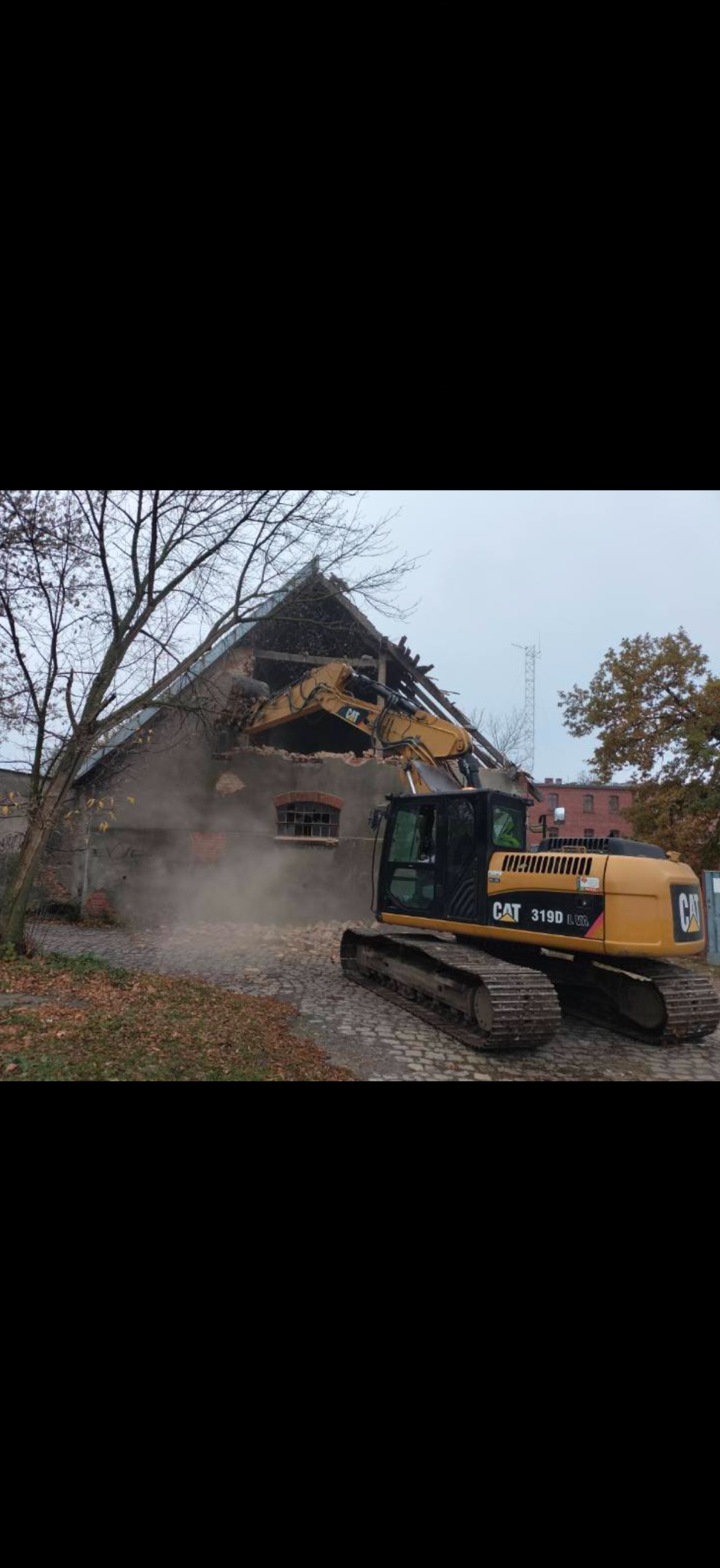 Żółta koparka CAT 319D L w trakcie rozbiórki starego, ceglanego budynku. Wokół gruz i pył, widoczne drzewa bez liści. Pochmurne niebo w tle.