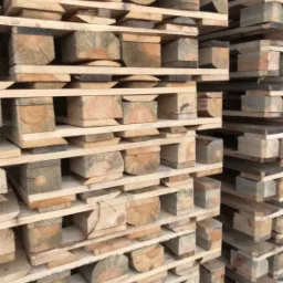 Stos ułożonych palet z jasnego drewna, widoczne przekroje belek z wyraźnymi słojami, konstrukcja palet z drewnianych desek i klocków.