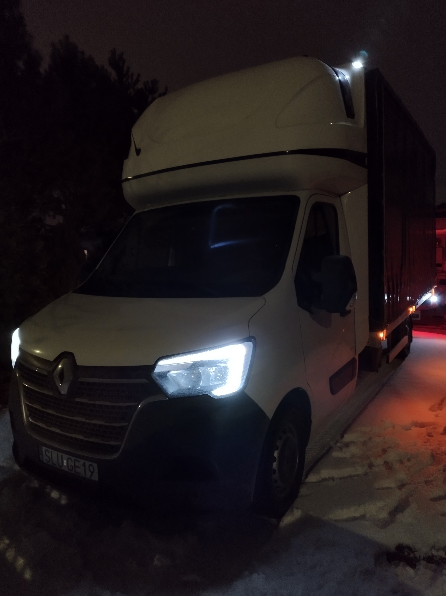 Biały Renault Master z wysoką zabudową i tablicą rejestracyjną SLU GE19, zaparkowany na śniegu w nocy, z włączonymi światłami.