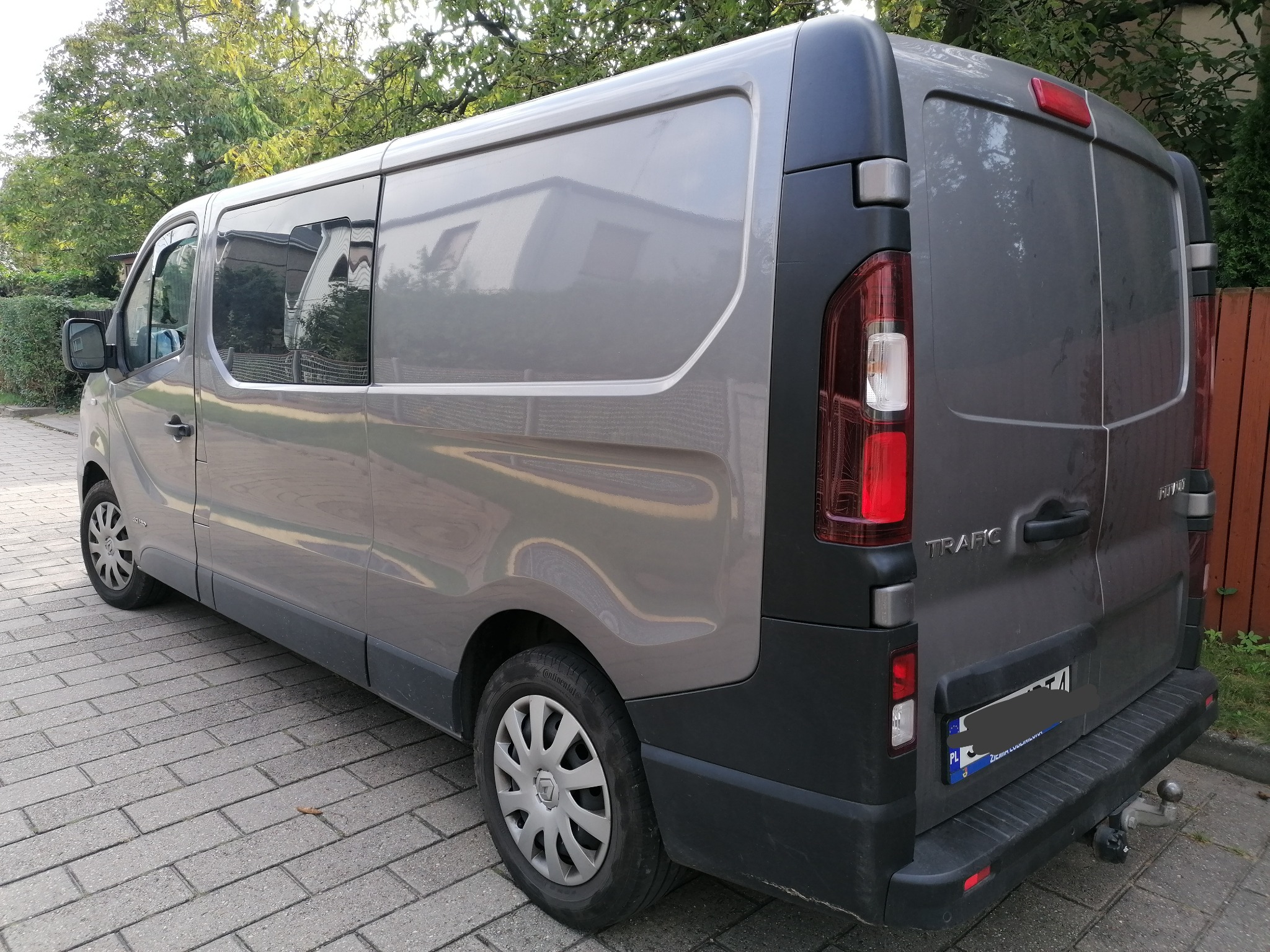 Szaro-grafitowy bus Renault Trafic z przyciemnianymi szybami, zaparkowany na brukowanej ulicy, widoczny z boku i tyłu, z zaczepem holowniczym.