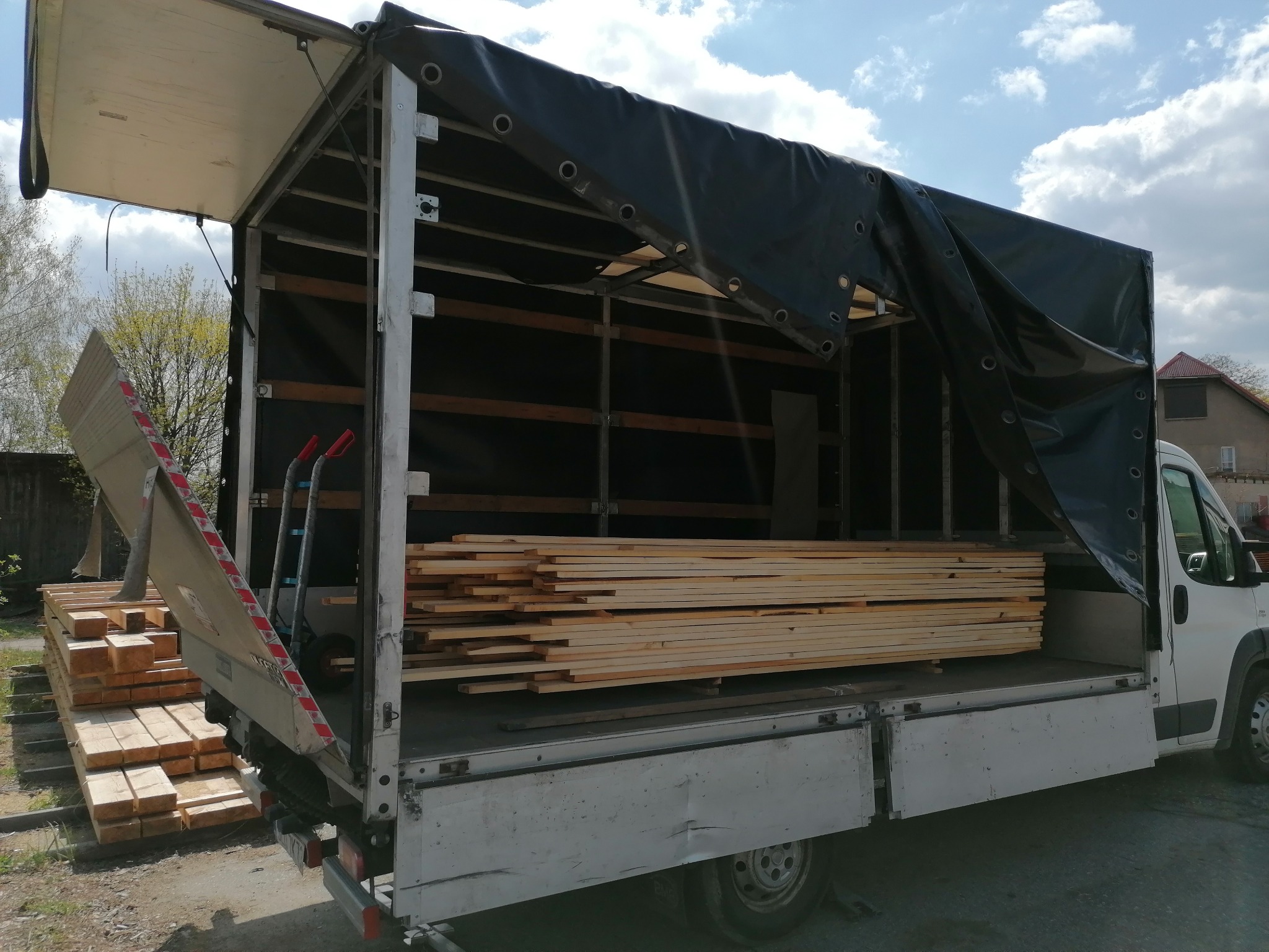 Częściowo rozładowany biały bus z plandeką, wypełniony drewnem budowlanym, z wózkiem transportowym w tle.